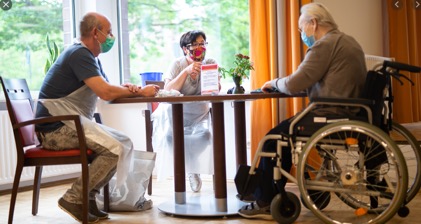 three masked people sitting at a table with one older man in a wheelchair