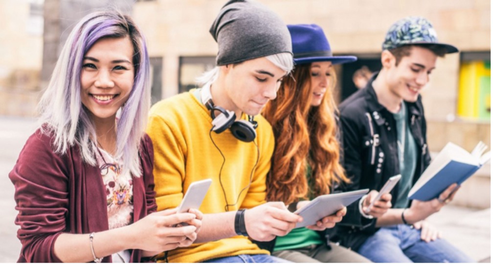four teenagers sitting on a bench 2 with iPhones one with iPad and one with book