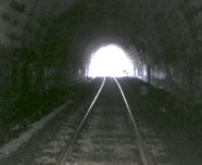 black and white photo of tunnel with train tracks and daylight at the end of the tunnel