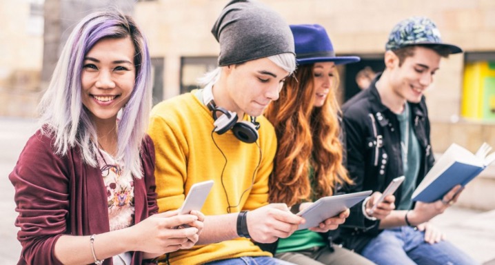 four teenagers sitting on a bench 2 with iPhones 1 with iPad and 1 reading a book